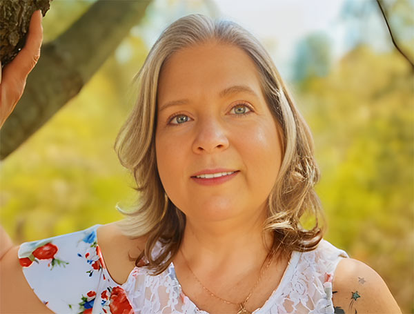 headshot of christina polnar in a white floral pattern dress, leaning against a tree